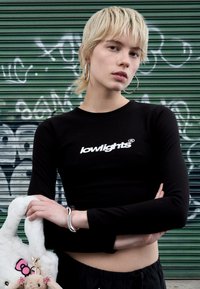 Black long-sleeve top with white "lowlights" logo, paired with a soft, white furry bag featuring cartoon accents, silver bracelet, against green backdrop.