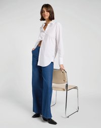 Camisa branca de manga longa com detalhes em botão, combinada com jeans azul de perna larga. Modelo está ao lado de uma cadeira bege com pernas em cromo.