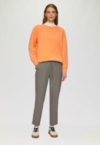 Oranje sweatshirt met reliëftekst "JOYFUL", grijze sweatpants met trekkoord en lichte sneakers in crèmekleurig en zwart. Soepele textuur, casual ontwerp.