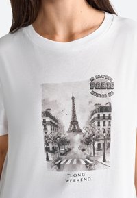 Hvid bomulds T-shirt med et monochromt print af Eiffeltårnet med omgivende bygninger og træer, teksten lyder "ER ALTID PARIS."