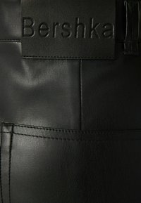 Svarta byxor i konstläder med en texturerad yta, präglad "Bershka"-logotyp på midjebandet, och synliga sömmar.