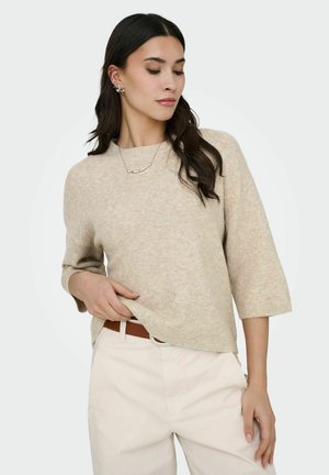 Žena s dlhými tmavými vlasmi, oblečená v béžovom krátkos rukávovom svetri, bielych nohaviciach, hnedom opasku, striebornej retiazke a kvetinových náušniciach, pozerá sa dole.