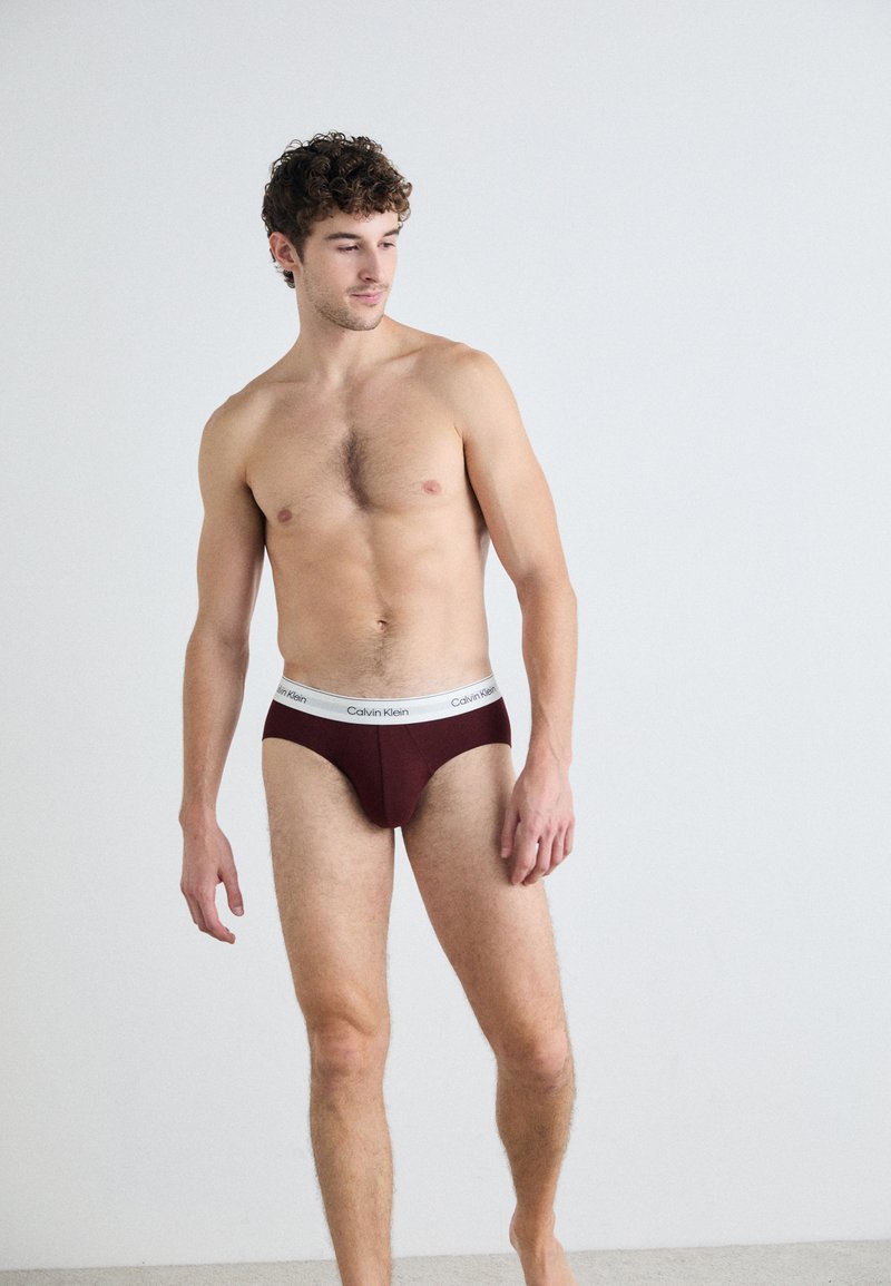 Bordó pamut alsónadrág fehér rugalmas derékkal, amelyen a Calvin Klein logo látható. A modell mezítláb áll egy semleges háttér előtt.