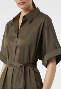 Olivengrünes Kleid mit einem Kragen-Halsauschnitt, kurzen hochgerollten Ärmeln und einem gebundenen Taillengürtel. Glattes Material mit einem leichten Glanz.