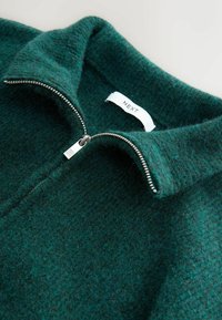 Pulover verde din lână cu guler cu fermoar. Dispune de un fermoar argintiu și o etichetă pe care scrie "NEXT". Textură moale cu un ușor model heather.