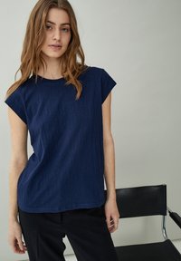 Top de color azul marino, de manga corta y corte relajado, con textura y escote redondo, combinado con pantalones oscuros y colocado sobre un fondo neutro.