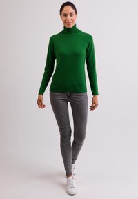 Zielony sweter z golfem wykonany z miękkiego materiału, dopasowany krój, długie rękawy, połączony z wąskimi szarymi dżinsami i białymi trampkami. Gładkie tło.
