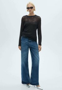 Maglione nero a maniche lunghe con trama aperta, abbinato a jeans blu a gamba larga e scarpe argento a punta. Design moderno e minimalista.
