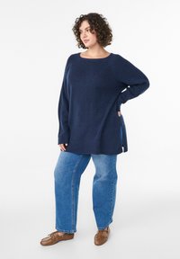 Maglione blu navy lavorato a maglia con scollo rotondo, maniche lunghe, spacchi laterali e motivo strutturato, abbinato a jeans larghi azzurri e scarpe marroni.