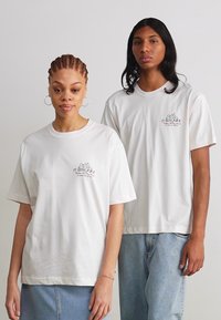 Camisetas de algodón blancas con cuello redondo y mangas cortas. La parte frontal presenta una impresión gráfica en verde y negro, combinada con jeans azules holgados.