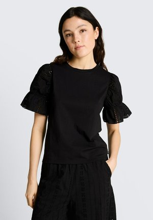 Młoda kobieta o długich ciemnych włosach nosi czarny top z bufkami i rękawami z ażurowej koronki oraz pasujące czarne spodnie z ażurowej koronki, stojąc na jednolitym tle.