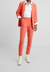 Oranžový oblek na míru se sakem na jedno knoflíkové zapínání, štíhlými kalhotami a bílou proužkovanou košilí. Obsahuje strukturovanou látku a krémové kotníkové boty.
