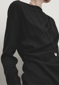 Black long-sleeved top with a smooth, textured fabric featuring gathering detail on the side. Includes a thin black necklace with a silver pendant.