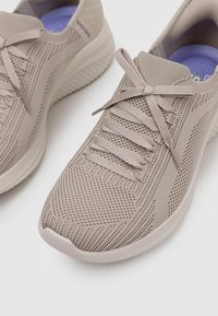 Scarpe sportive grigio chiaro con una tomaia in maglia traspirante, lacci piatti, suola ammortizzata e dettagli testurizzati sottili. Fodera interna viola visibile.