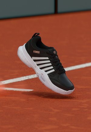 Melnbalts K-Swiss sporta apavu pāris, kas iespēra sarkanā māla virsmā tenisa kortā pie baltas robežlīnijas.