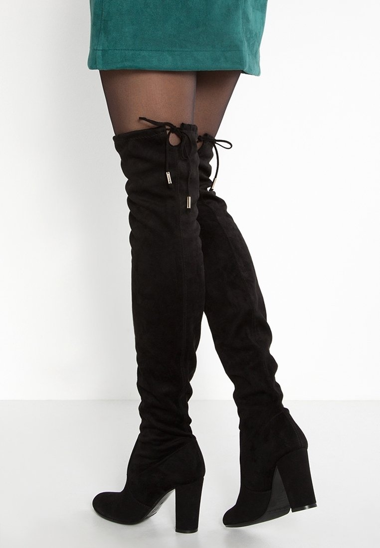 Mujer vestida con medias negras transparentes, una falda verde y botas de tacón alto de ante negro hasta el muslo con cintas traseras, sobre un fondo blanco.