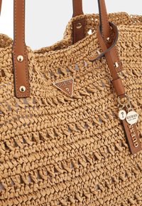 Bolso tote tejido marrón hecho de fibras naturales, con asas de cuero sintético marrón y un logo de marca metálico. Patrón de tejido abierto texturizado.