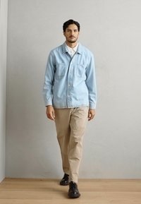 Light blue denim shirt with two chest pockets, beige trousers, and black shoes. Model stands upright against a plain background.