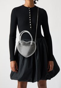 Stříbrná kabelka ve tvaru srdce s dvojitými uchy a řetízkovým popruhem, vyrobená z texturovaného metalického materiálu, nošená s černými žebrovanými šaty a sukní.