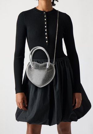 Sølv hjerteformet taske med dobbelte håndtag og kædestrop, lavet af et struktureret metallisk materiale, båret med en sort ribbad kjole og nederdel.