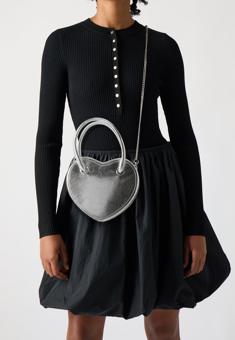 Stříbrná kabelka ve tvaru srdce s dvojitými uchy a řetízkovým popruhem, vyrobená z texturovaného metalického materiálu, nošená s černými žebrovanými šaty a sukní.