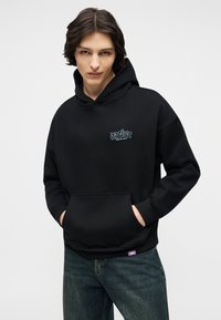 Mlajša oseba s srednje dolgimi temnimi lasmi, oblečena v črno kapuco z majhnim logotipom na prsih in modrimi kavbojkami, z rokami v žepu kapuce.