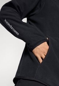 Pull en polaire noir avec col haut, poches latérales et logo "Peak Performance" brodé sur la manche. Texture douce et design fonctionnel.