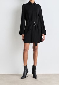 Donna che indossa un abito camicia nero a maniche lunghe con cintura, abbinato a stivaletti neri con punta a stiletto, in piedi davanti a una parete bianca uniforme.