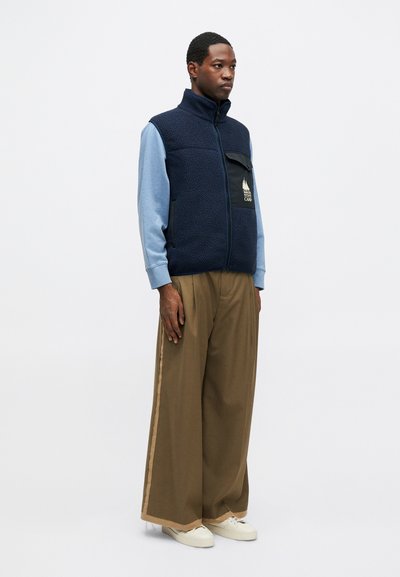 Námornícky fleecová vesta s vysokým golierom, modré dlhé rukávy, veľká kapsa a logo; v kombinácii s hnedými voľnými nohavicami a bielymi teniskami.