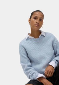 Jasnoniebieski sweter z dzianiny o prążkowanej strukturze, okrągłym dekolcie i opadających ramionach, noszony z białą koszulą z kołnierzykiem pod spodem.