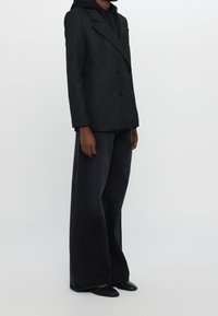 Blazer doppiopetto a quadri neri sopra una felpa con cappuccio nera, abbinato a jeans neri a gamba larga e scarpe nere flat, vista laterale.