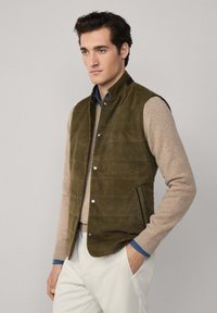 Olivově zelená sukně z nubuku s vysokým límcem, zapínáním na patentky a bočními kapsami, vrstvená přes krémový svetr s modrými akcenty.