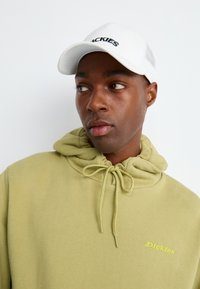 Giovane che guarda di lato, indossando un felpa leggera verde con cappuccio e un cappellino da baseball bianco con testo nero.