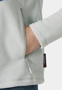 Chaqueta de forro polar azul claro con un bolsillo lateral con cremallera, que presenta una superficie texturizada y una etiqueta POLARTEC® en la costura lateral.