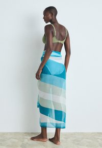Bufanda ligera y translúcida de tela en ondas azul y verde, drapeada hasta la mitad de la pantorrilla. La modelo lleva un sujetador de bikini verde oliva y está descalza.