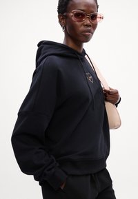 Črna kapuca z vrvico, ki ima logotip srca v roza zlati barvi. Model nosi roza sončna očala in drži svetlo roza torbico. Gladka tekstura.