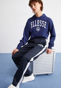 Jūras spēku kapuce ar "ELLESSE" logotipu, apvienota ar tumši pelēkām sporta biksēm ar baltām sānu svītrām, uz zila fona.