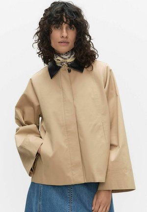 Kobieta o kręconych włosach, ubrana w beżową kurtkę z czarnym kołnierzem, z rękami w kieszeniach, niebieską jeansową spódnicę i wzorzystą apaszkę na szyi.