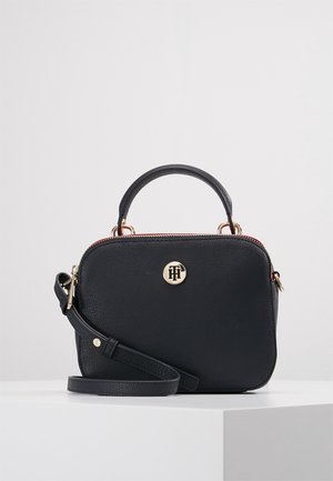 Sac bandoulière en cuir noir avec poignée supérieure, emblème logo doré et bandoulière réglable reposant sur une surface blanche contre un fond neutre.