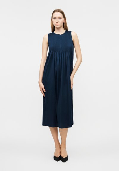 Γυναίκα που φοράει μίντι φόρεμα χωρίς μανίκια σε ναυτικό μπλε χρώμα με πλισέ μπούστο, στέκεται με το ένα χέρι στην τσέπη και μαύρα μυτερά παπούτσια.