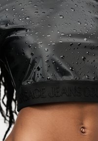 Μαύρο κομμένο τοπ με σταγόνες νερού και κεντημένο το "Versace Jeans Couture" στη μπάντα στο κάτω μέρος, φορεμένο από άτομο με ορατή κοιλιά.