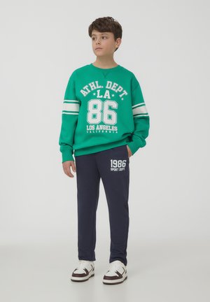 Felpa verde con grafiche bianche e maniche a righe, abbinata a pantaloni della tuta blu navy con logo stampato. Indossata con sneakers bianche e marroni.