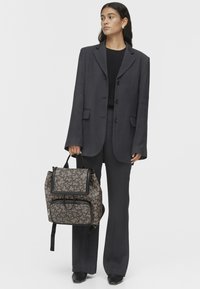 Zaino realizzato in tessuto stampato marrone e nero, con pattina superiore, tasca frontale con zip e spallacci regolabili.