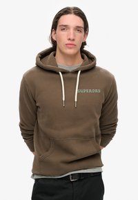 Sudadera marrón con bolsillo frontal, capucha con cordón y detalles en blanco que presentan el logotipo "SUPERDRY" en bordado azul. Usada sobre una camiseta gris.