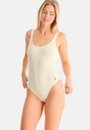 Femme blonde portant un body crème sans manches, debout avec un léger sourire sur un fond blanc uni.