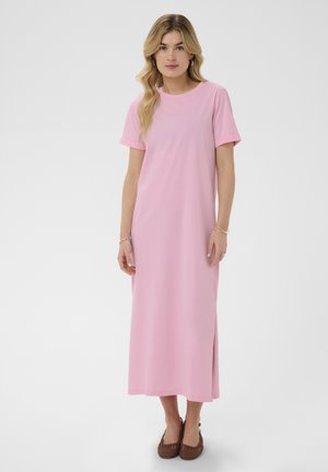 Frau mit langen blonden Haaren, die ein hellrosa Maxikleid mit kurzen Ärmeln und braune geflochtene Ballerinas auf einem einfarbigen hellen Hintergrund trägt.