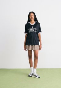 Svart Nike-trøye med hvitt logo og kant, kombinert med et beige skjørt. Modellen har på seg hvite sokker og svarte joggesko. Minimalistisk bakgrunn.
