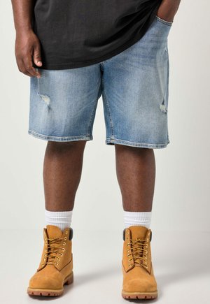 Person iført lyseblå revnede denimshorts, hvide sokker og beige arbejdstøvler mod en ensfarvet lys baggrund.