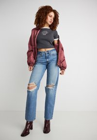 Νέα γυναίκα με σγουρά μαλλιά, φορώντας ένα κοντό μαύρο μπλουζάκι με γραφικό σχέδιο, μπουφάν μπορντό, σκισμένο μπλε τζιν και μπορντό μποτάκια στον αστράγαλο, στέκεται μπροστά σε έναν απλό τοίχο.