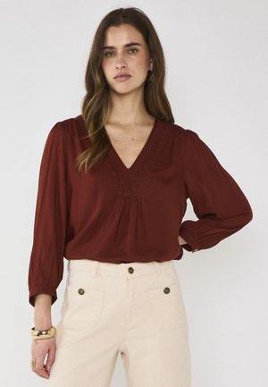 Femme portant un chemisier à col en V couleur rouille avec manches longues et un pantalon crème taille haute, debout avec une main sur la hanche, sur fond blanc.